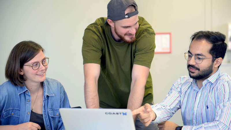 Three people are sitting in front of laptop, one is talking and pointing at the screen.