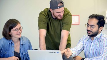 Three people are sitting in front of laptop, one is talking and pointing at the screen.